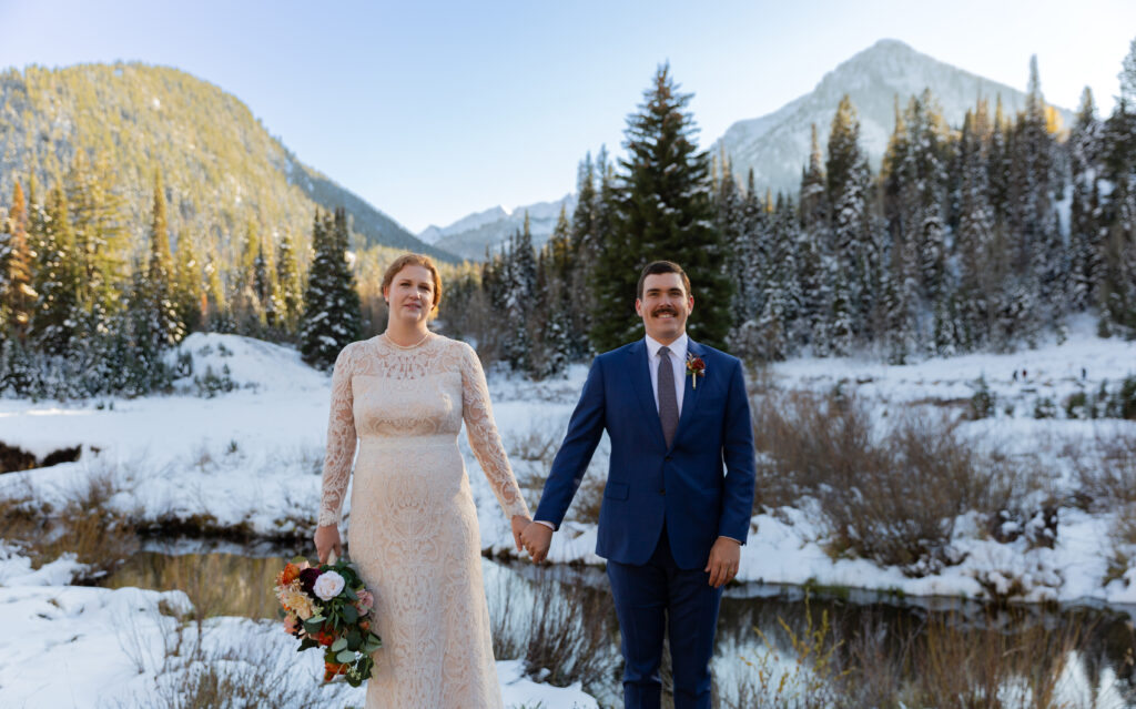 Bride and groom standing apart facing the camera and holding hands in front of the snowy mountains in Salt Lake City, Utah