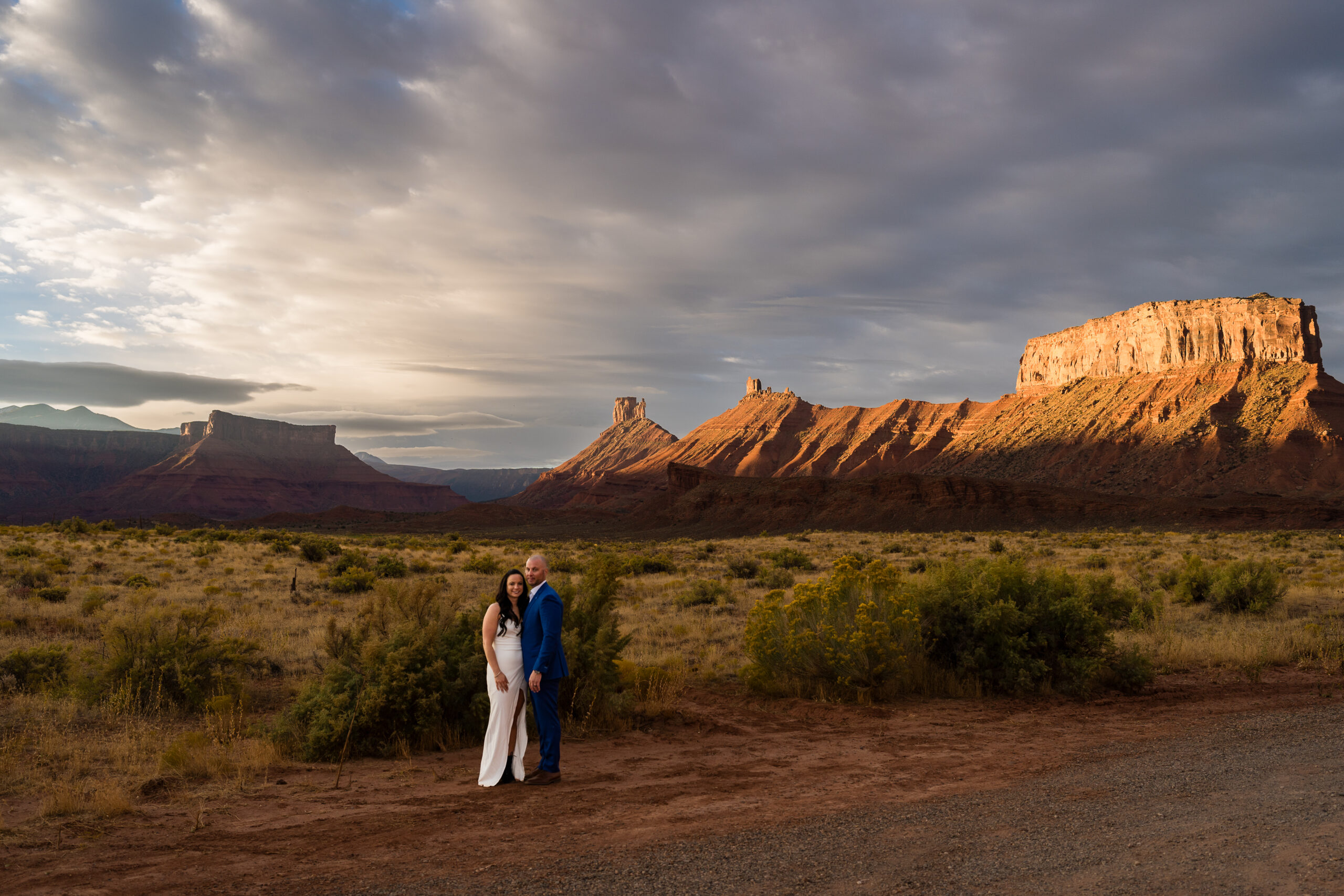 Bride and groom standing together in an embrace looking at the camera in front of the buttes in Castle Valley at sunrise in Moab, Utah
