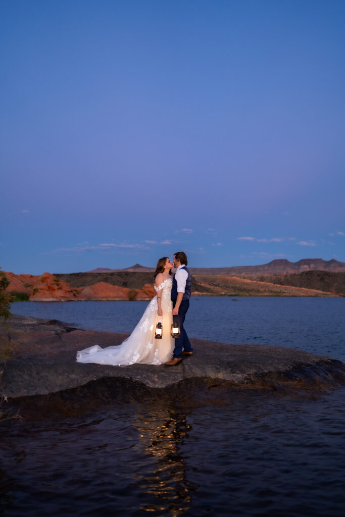 Bride and groom facing each other and kissing on the red rock shore by the water while holding lanterns in their hands