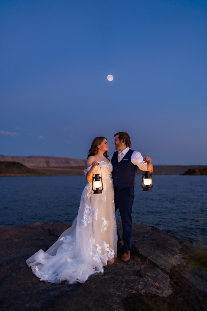 Bride and groom facing each other and looking at one another while holding their lanterns up on the red rock shore by the water under the moonlight