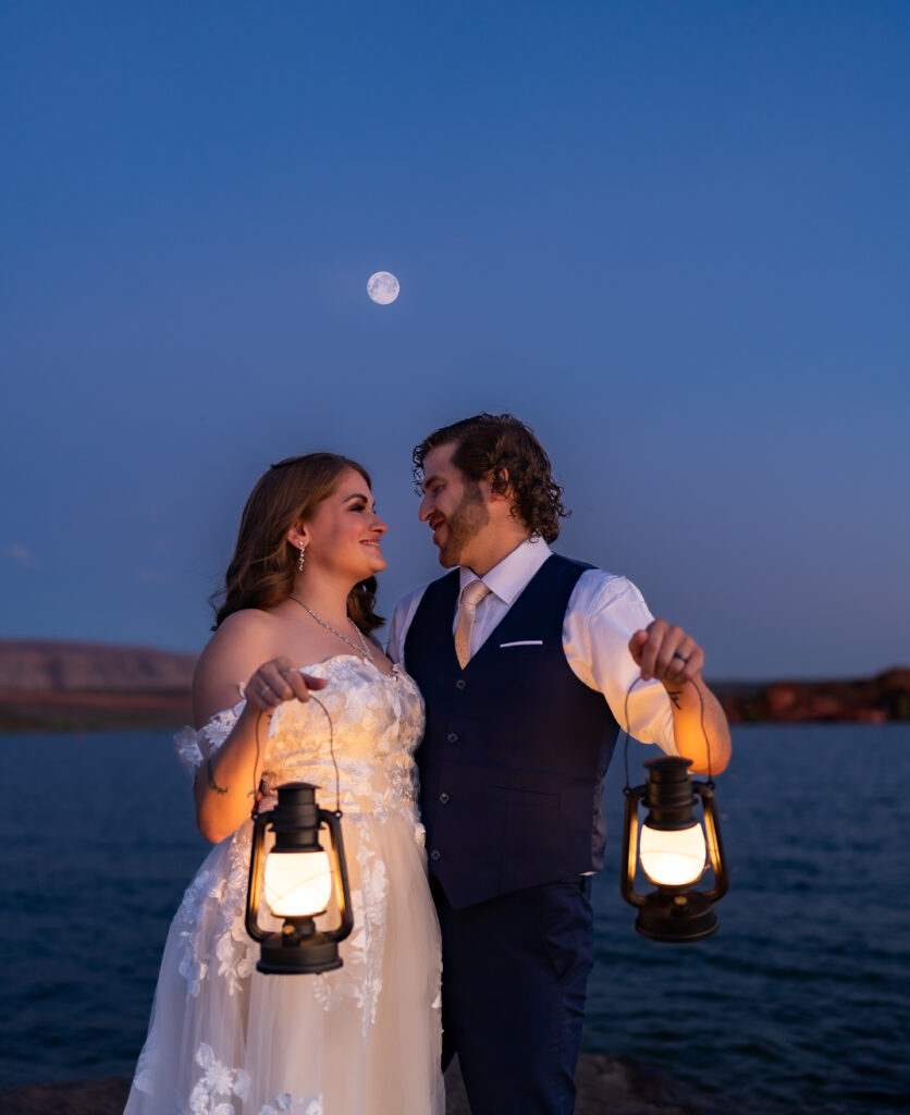 Bride and groom facing each other and looking at one another while holding their lanterns up on the red rock shore by the water under the moonlight