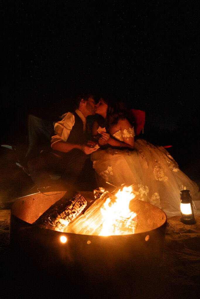 Bride and groom kissing in while sitting by the campfire and holding s'mores