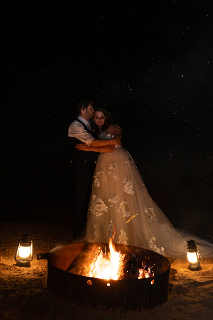 Bride and groom standing by campfire while groom kisses the side of bride's head