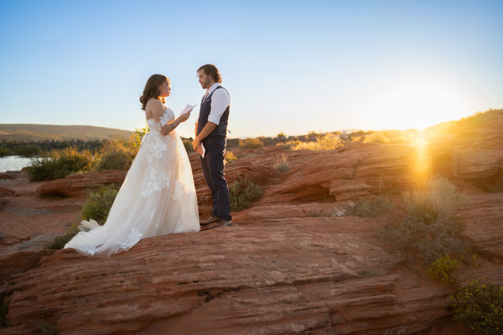 Bride and groom standing on red rock cliff reading vows o each other at sunset