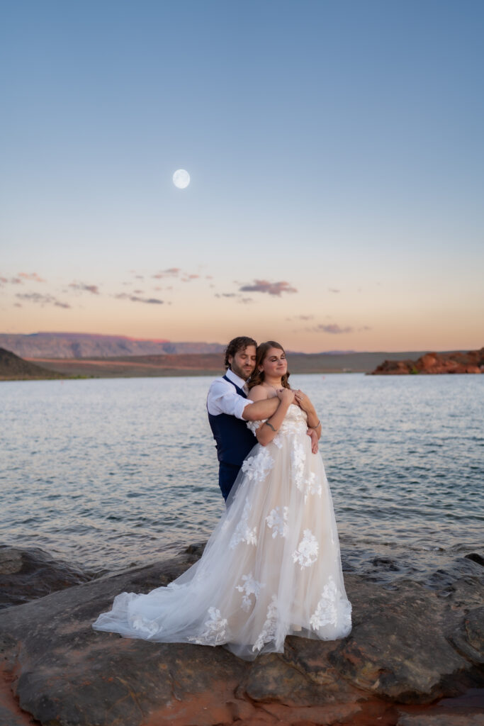 Groom standing behind bride with his arms around her on the red rock cliffs by the water as the sun is setting and the moon is rising