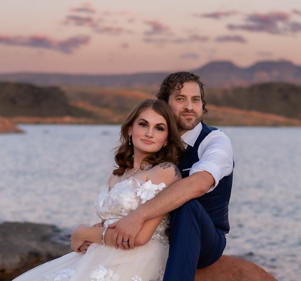 Bride and groom sitting on red rock shore by the water looking at the camera