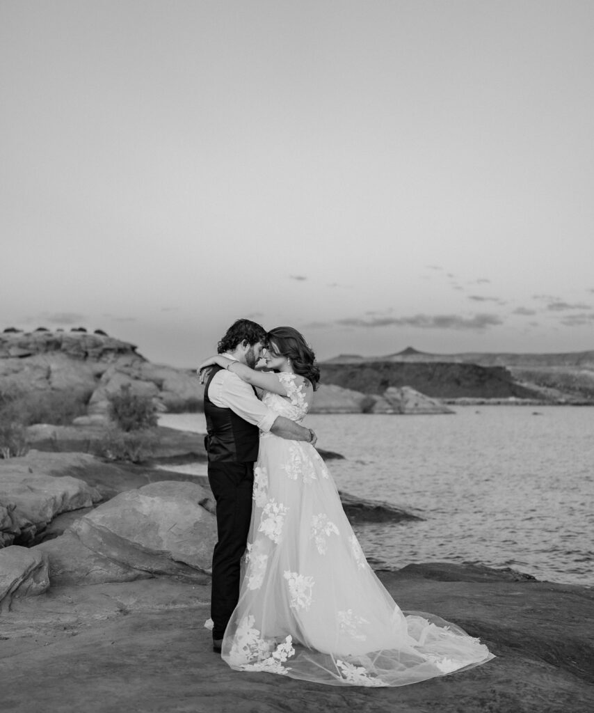 Black and white photo of bride and groom facing each other with their arms around each other and touching foreheads on the red rock shore near the water