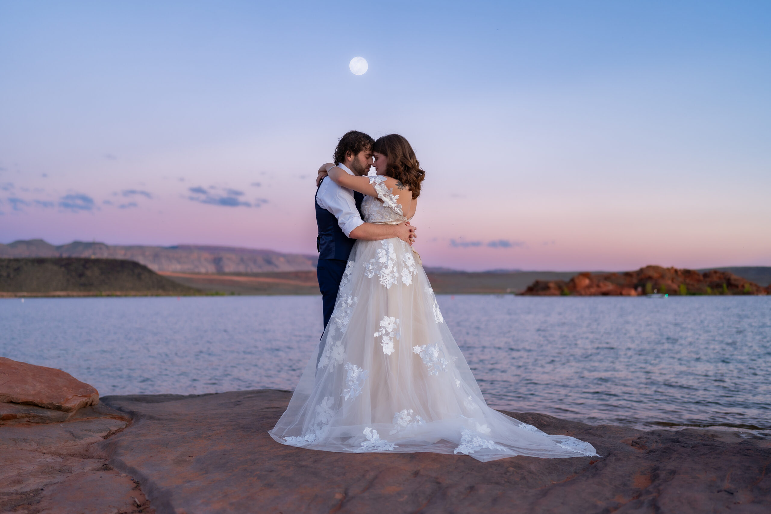 Bride and groom facing each other with their arms around each other and touching foreheads on the red rock shore near the water