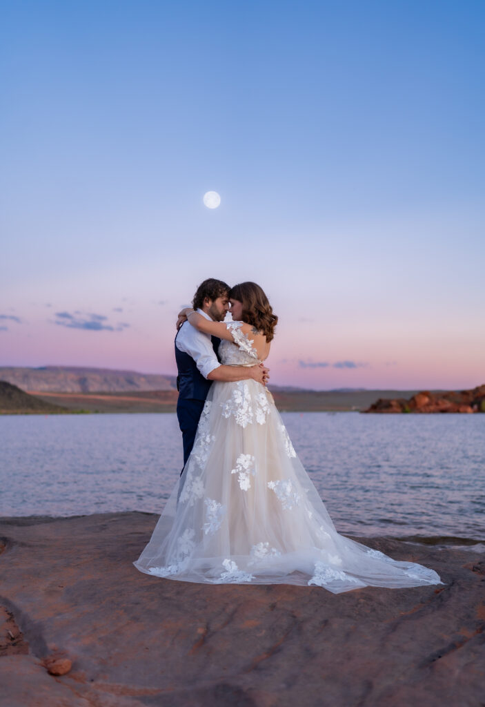 Bride and groom facing each other with their arms around each other and touching foreheads on the red rock shore near the water with the moon in the background