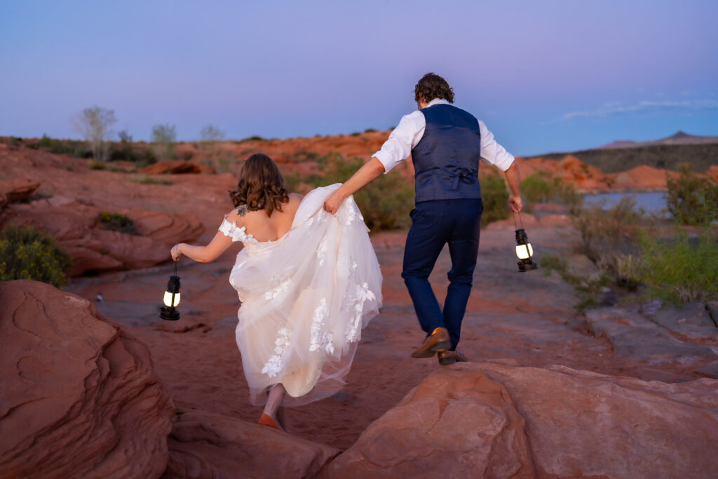 Bride and groom walking over red rocks holding lanterns and groom helping bride with her dress
